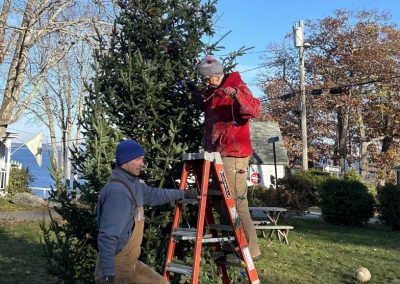 2023-11-14 Meri Elves, photographed in Bayside, ME by David Leaming. “Merithew Park residents erected and strung Christmas lights on a donated fir tree in the park in Bayside on Tuesday, November 14, 2023. Andrew Bewsher assists Brynna Ledyard with lights as canine helpers Yoko, left, and Blue watch. There will be a tree lighting ceremony on November 24 at 5:30 PM, and all are welcome.” #elves #christmastree #treelighting #merithewsquare #baysidemaine See less